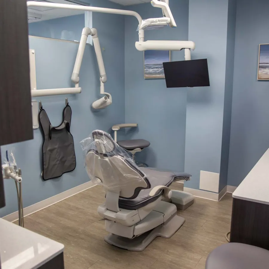 Modern dental office with patient chair, wall-mounted TV, X-ray equipment, and light blue walls.