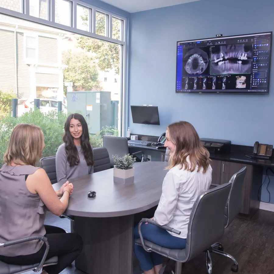 Three women in a modern office discussing dental X-rays displayed on a screen during a consultation meeting.