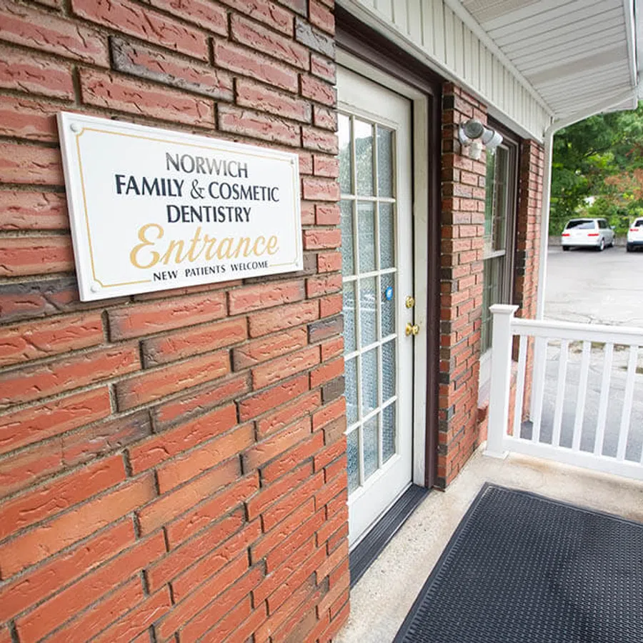 Entrance of Norwich Family & Cosmetic Dentistry with brick wall, white door, and parking lot in background