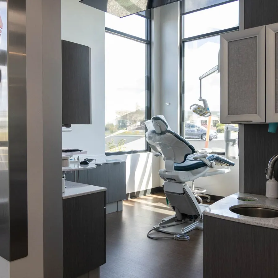 Modern dental office room with dental chair, large windows, cabinetry, and sink under natural light