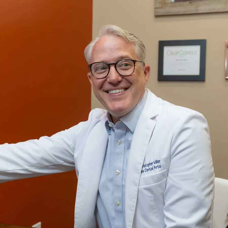 Smiling male dentist in white coat talking to a female patient in a dental office with certificates on the wall.