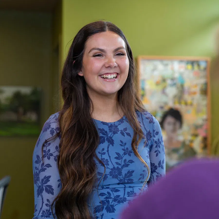 Smiling woman with long brown hair wearing a blue floral dress engaged in conversation indoors.