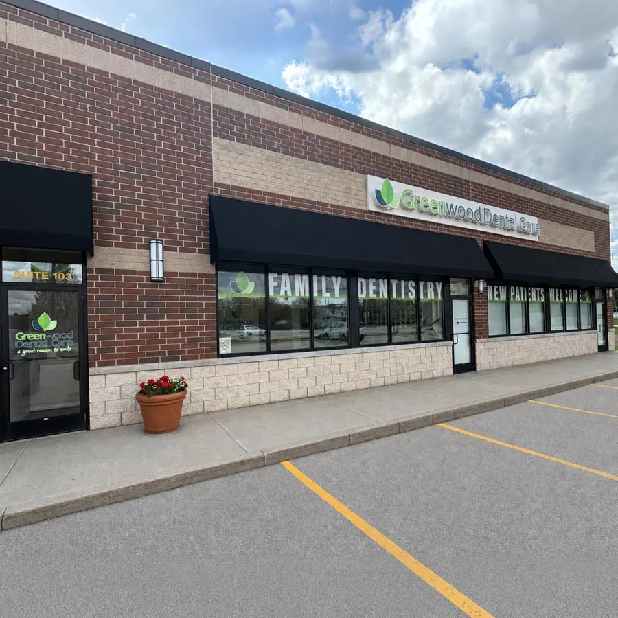 Exterior of Greenwood Dental Care with large windows, black awnings, and a parking lot on a partly cloudy day.