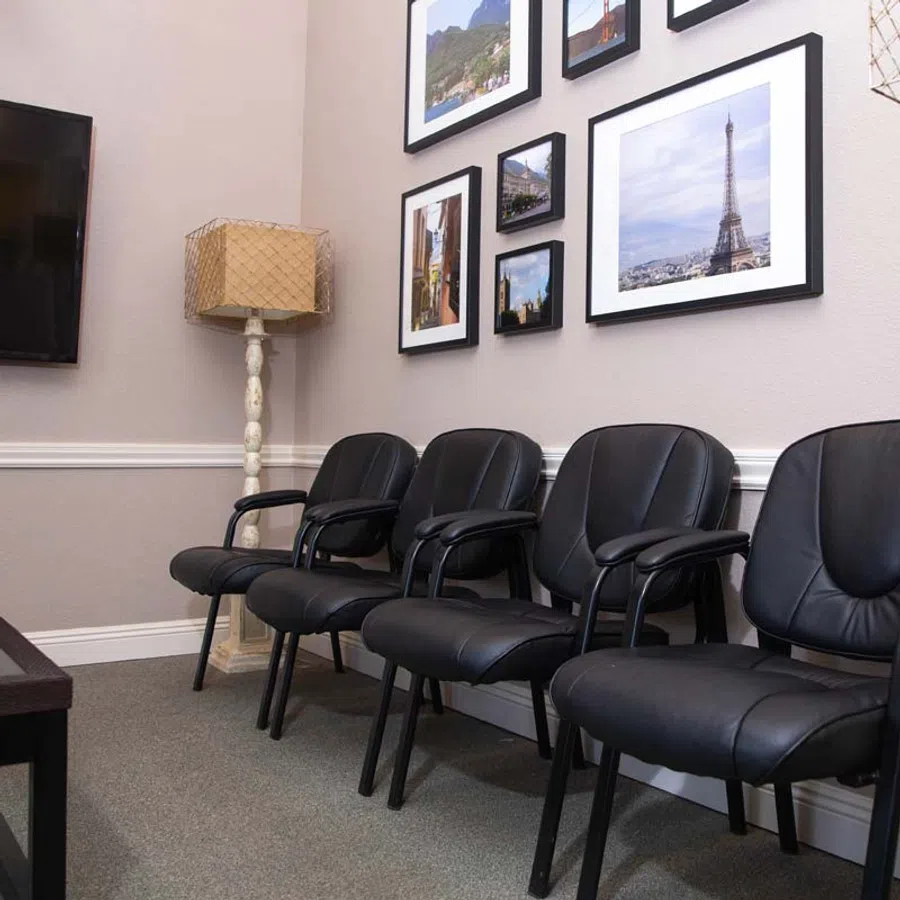 Row of black leather chairs in a waiting room with framed travel photos and a beige lamp on a white stand.