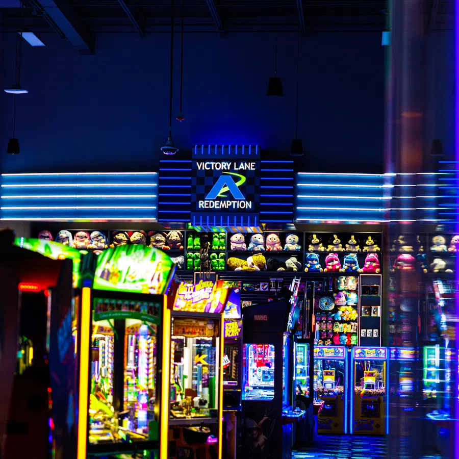 Brightly lit arcade redemption game area with plush toys and colorful machines under Victory Lane sign.