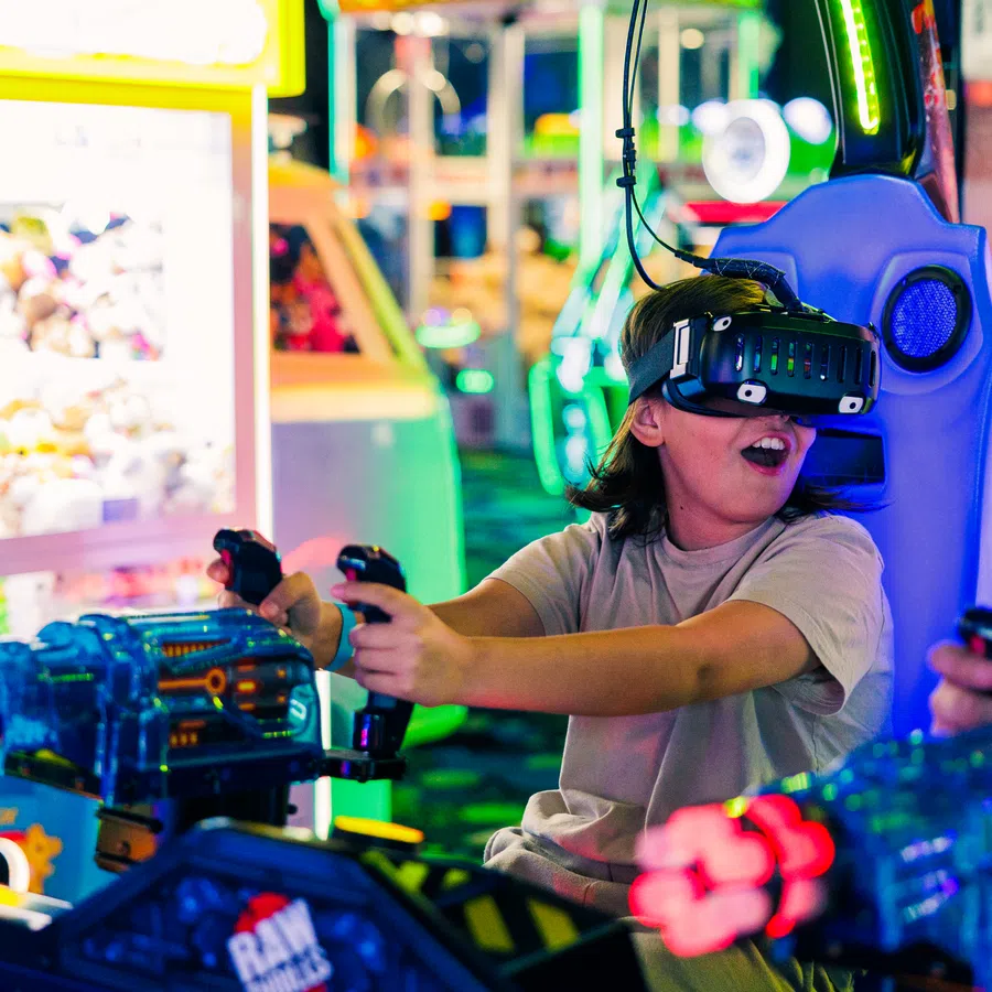 Child wearing VR headset playing an arcade shooting game with bright neon lights and colorful machines.