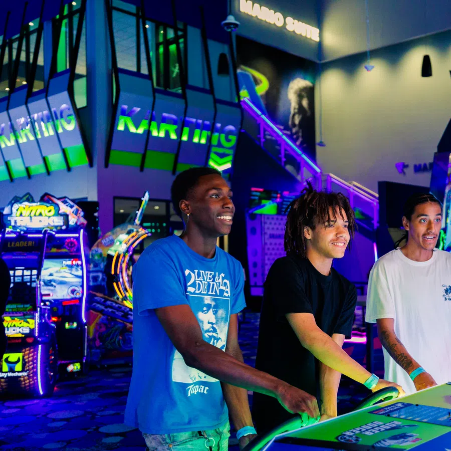 Three young men enjoying arcade games with vibrant neon lights and colorful gaming machines in the background.