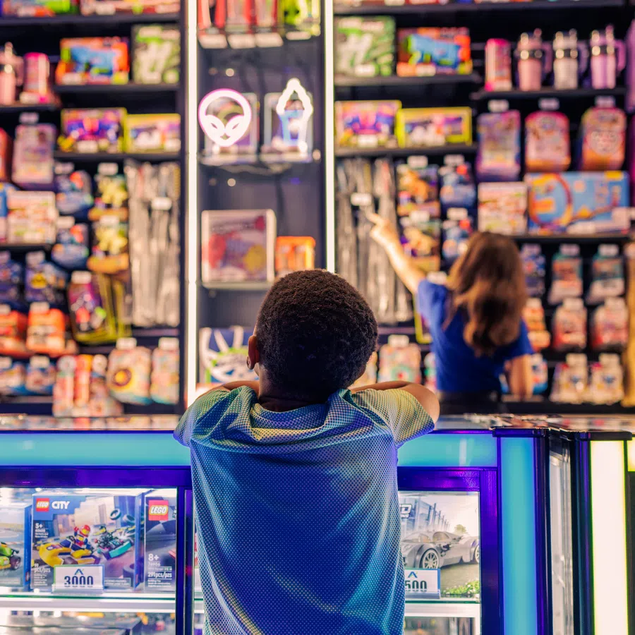 Children browsing colorful toys and prizes at a carnival game booth with glowing blue and white display cases.