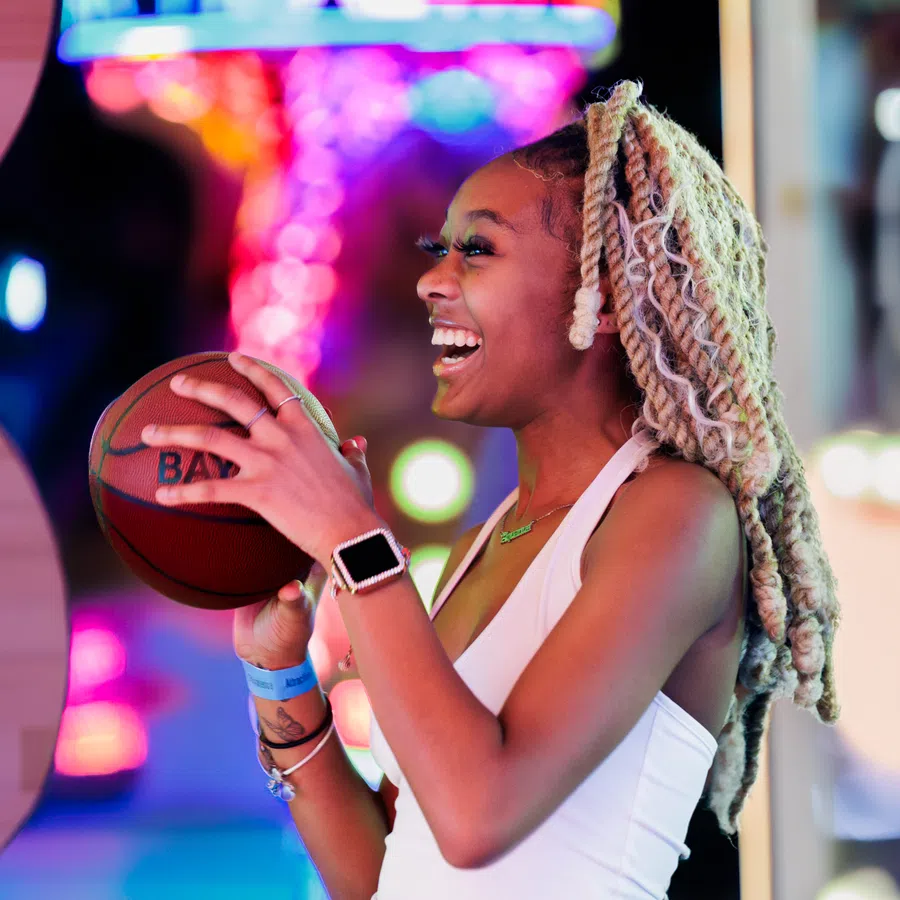 Young woman with blond twists laughing while holding a basketball at a vibrant, colorful arcade game.