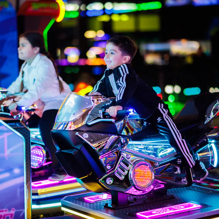 Two children enjoying a glowing motorcycle arcade game with vibrant neon lights in a gaming arcade.