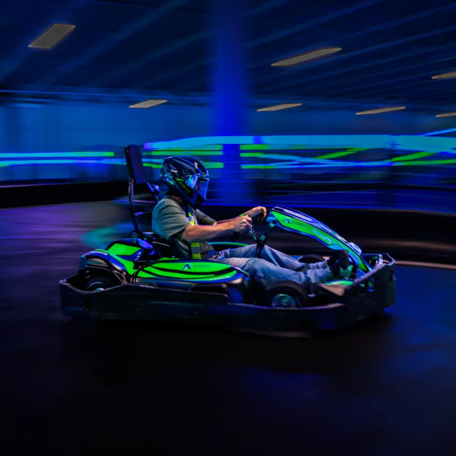 Person driving a green go-kart at high speed on an indoor track with motion blur and neon lights.
