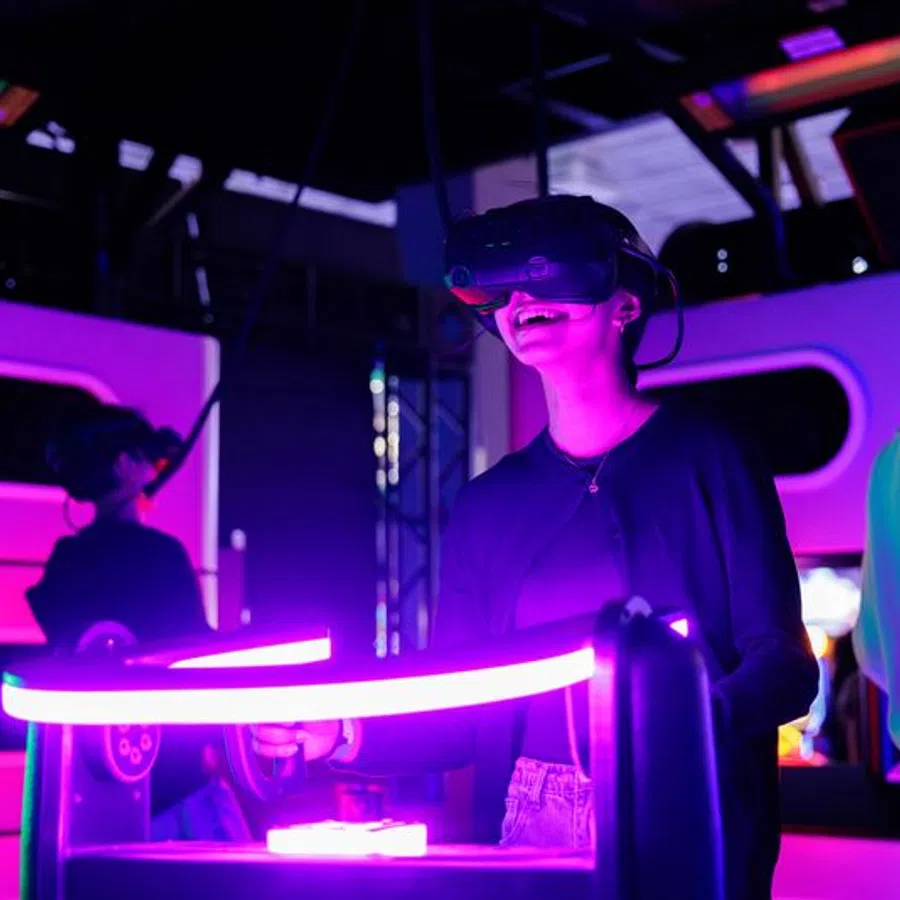 Person smiling while using virtual reality headset and controllers in neon-lit gaming arcade.