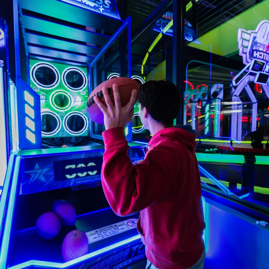 Person in a red hoodie playing a basketball arcade game in a neon-lit entertainment center.