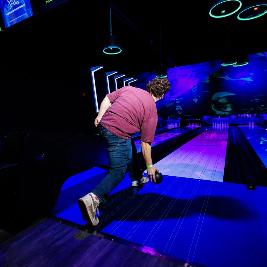 Man wearing casual clothes bowling under neon lights at an indoor bowling alley with digital scoreboards.