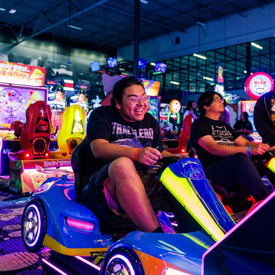 Three friends enjoy racing arcade games in a colorful, neon-lit arcade with various game machines in the background.