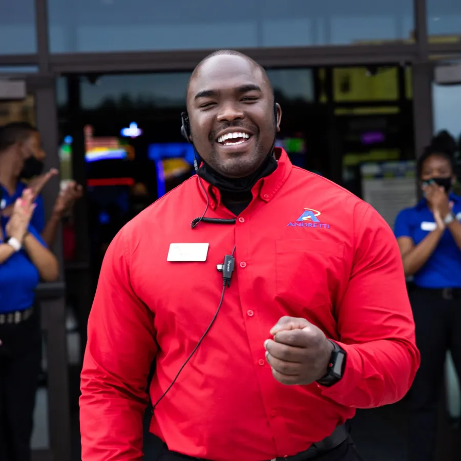 Smiling employee in red shirt welcomes guests outside, with team members in blue shirts clapping.