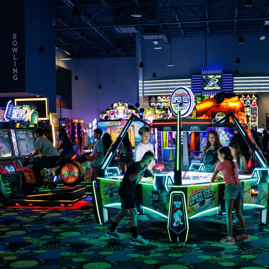 Children and teens playing air hockey and arcade games in a vibrant, neon-lit indoor arcade center.