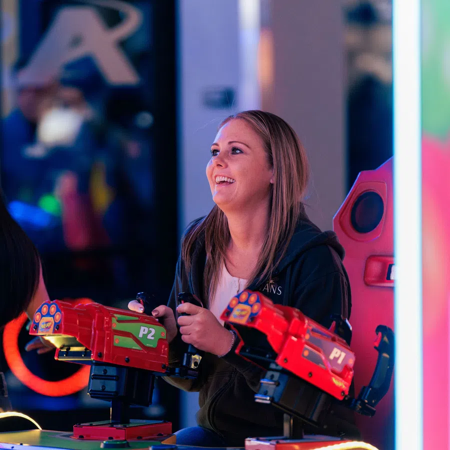 Smiling woman playing a red arcade shooting game with dual guns in a vibrant gaming environment.