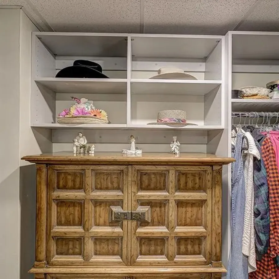 Wooden cabinet in a walk-in closet with shelves holding hats, figurines, and hanging clothes including colorful shirts and jackets.