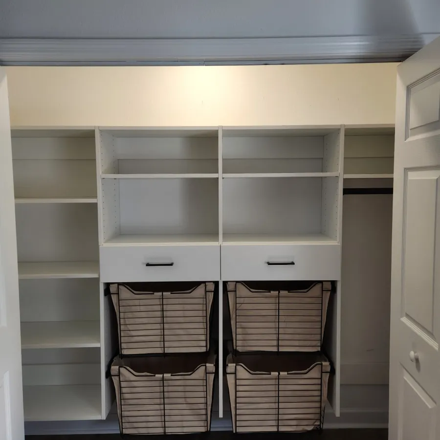 Empty white built-in closet organizer with shelves, drawers, and fabric baskets on wood floor in bright room.