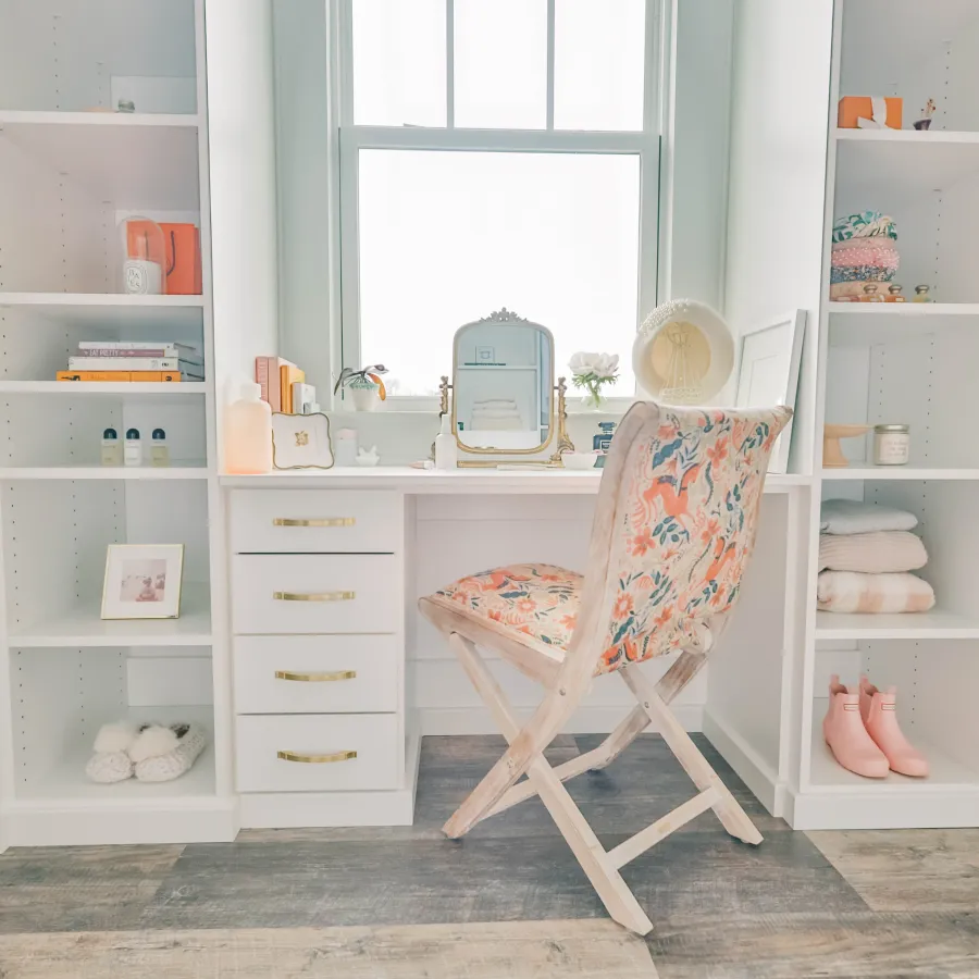 Walk in closet with white shelves, glass doors, white vanity area with floral cushioned chair, a mirror, hanging clothes in stylish dressing room