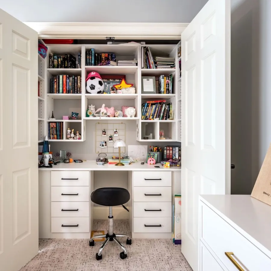 Home office closet with white shelves, desk, black swivel chair, books, and decorative items inside open double doors.
