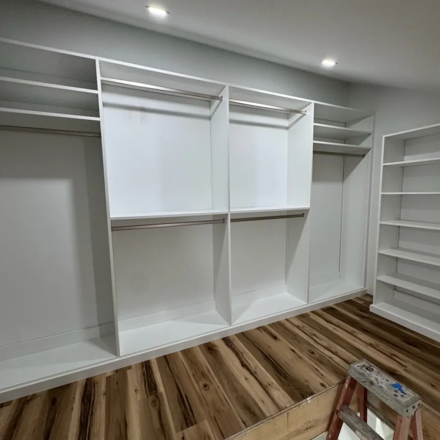 Empty white built-in closet with multiple shelves and hanging rods, wooden floor, and ladder in an unfinished room.