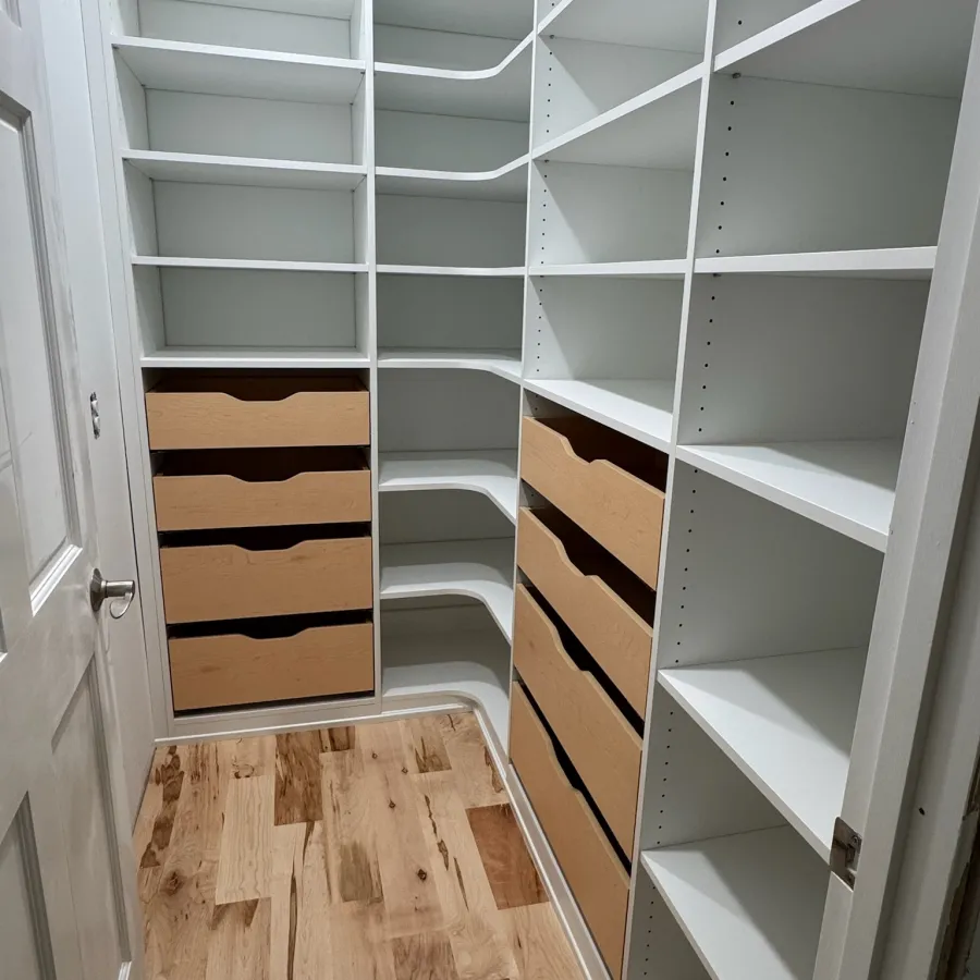 Walk-in closet with white shelves, built-in wooden drawers, and light hardwood flooring in a narrow room.
