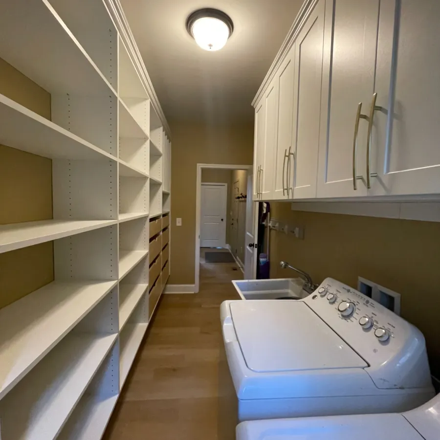 Laundry room with white cabinets, open shelving, washer and dryer, and beige walls and flooring.