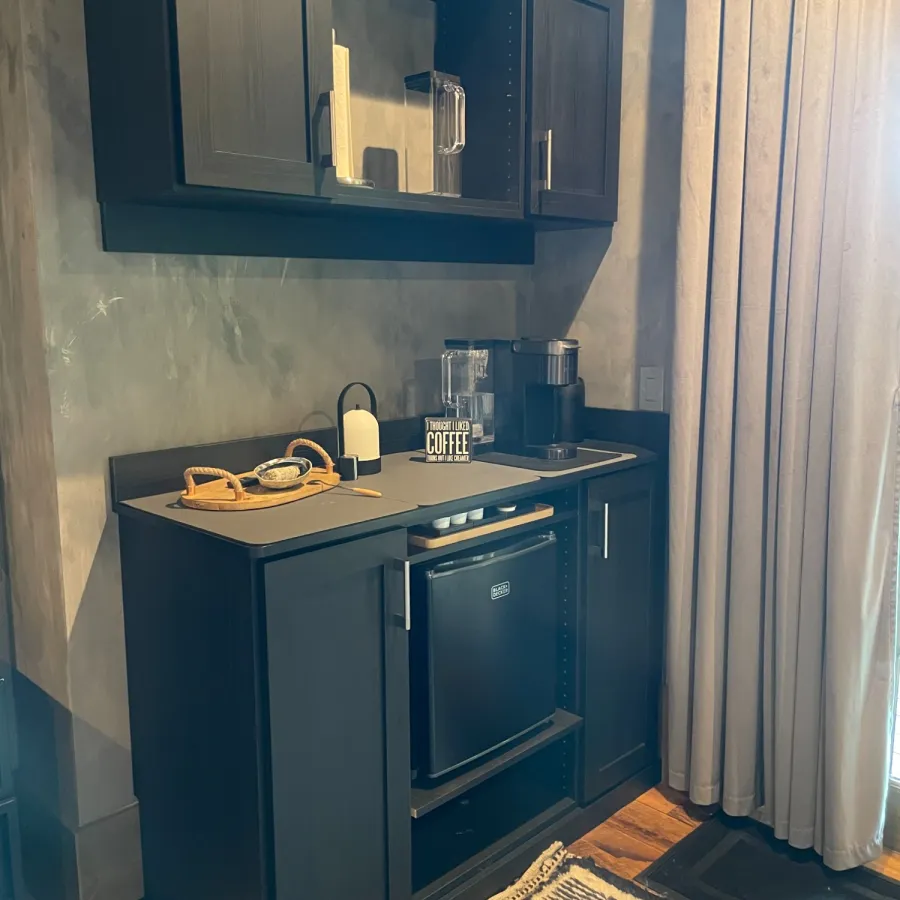 Compact black mini kitchen with cabinets, mini fridge, coffee maker, and neatly arranged accessories on wood floor.