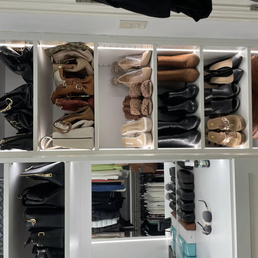 Organized closet shelves with various handbags, boots, heels, and shoes in neutral and black tones under bright lighting