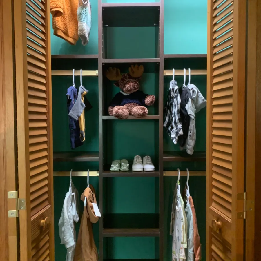 Organized closet with wooden shelves, hanging baby clothes, shoes, hats, and soft toys against a teal wall.