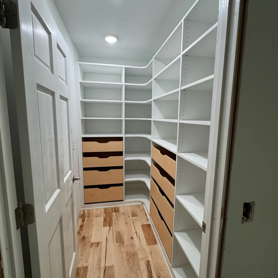 Spacious walk-in closet with white shelves, wood drawers, and hardwood floor under ceiling lights.