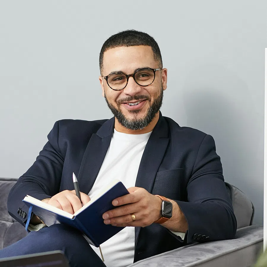 Smiling man in glasses and blazer writing in notebook while sitting on gray sofa in modern office.