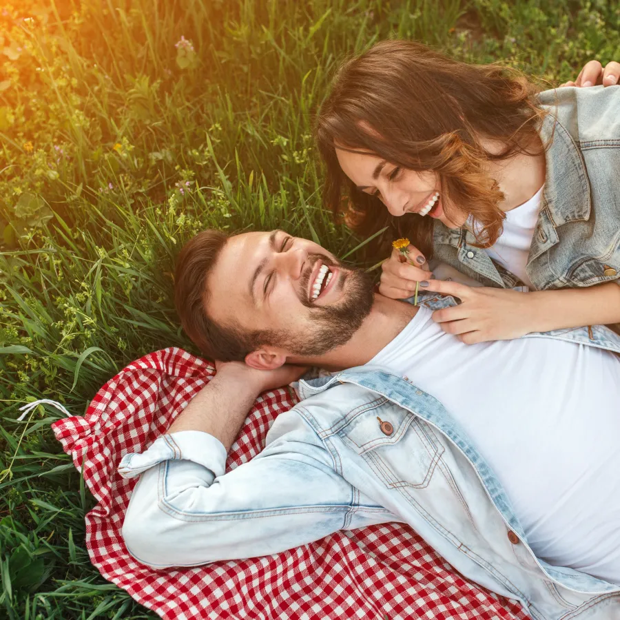 Couple laughing and relaxing on a red checkered blanket in a sunny green park.
