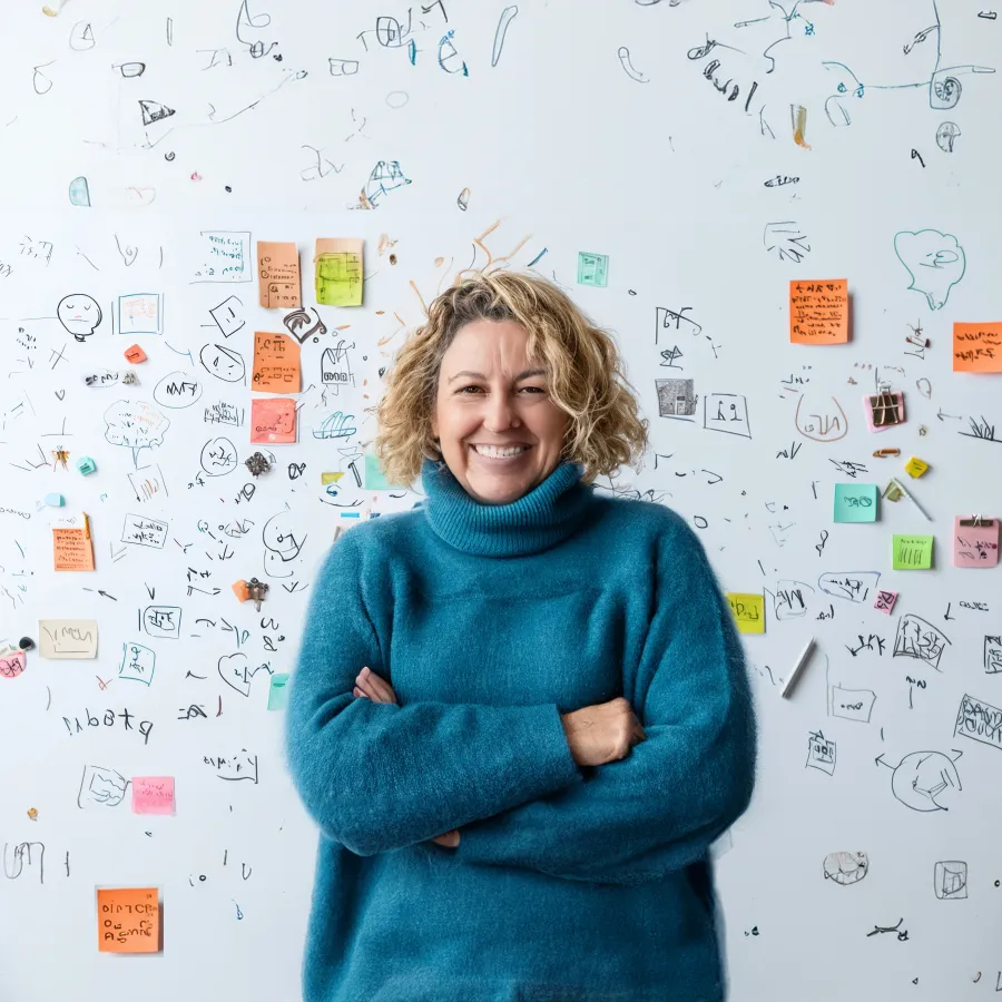 Smiling woman in blue sweater standing with arms crossed in front of whiteboard with notes and sketches.