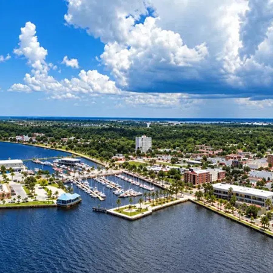 Aerial view of a coastal city with marina, residential buildings, greenery, and cloudy blue sky.