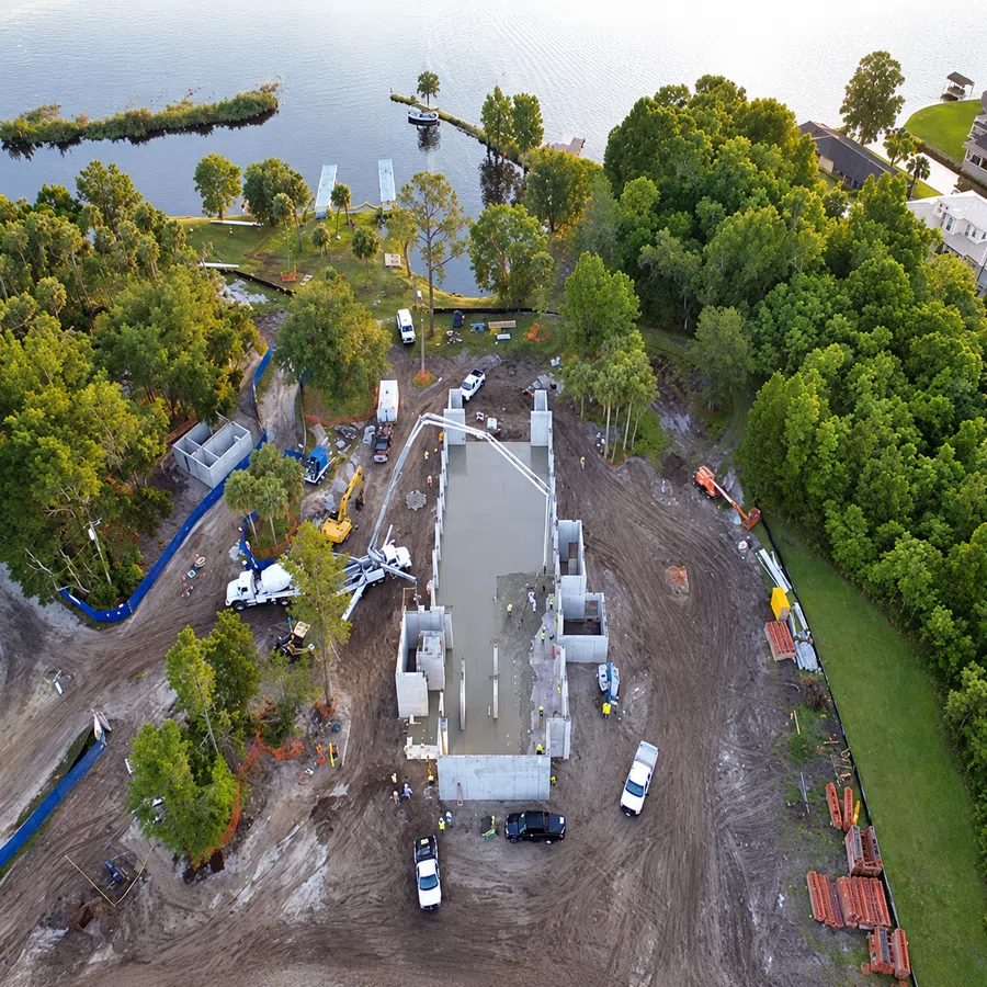 Aerial view of residential construction site near a lake with concrete foundation and surrounding trees.