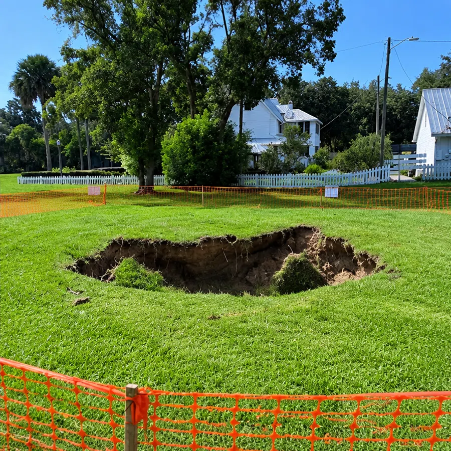Large sinkhole in grassy area surrounded by orange safety fencing near white houses and trees.
