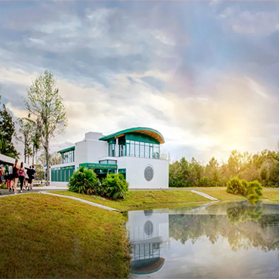 Modern white building with green roof by a reflective pond and people walking outdoors on a cloudy day.