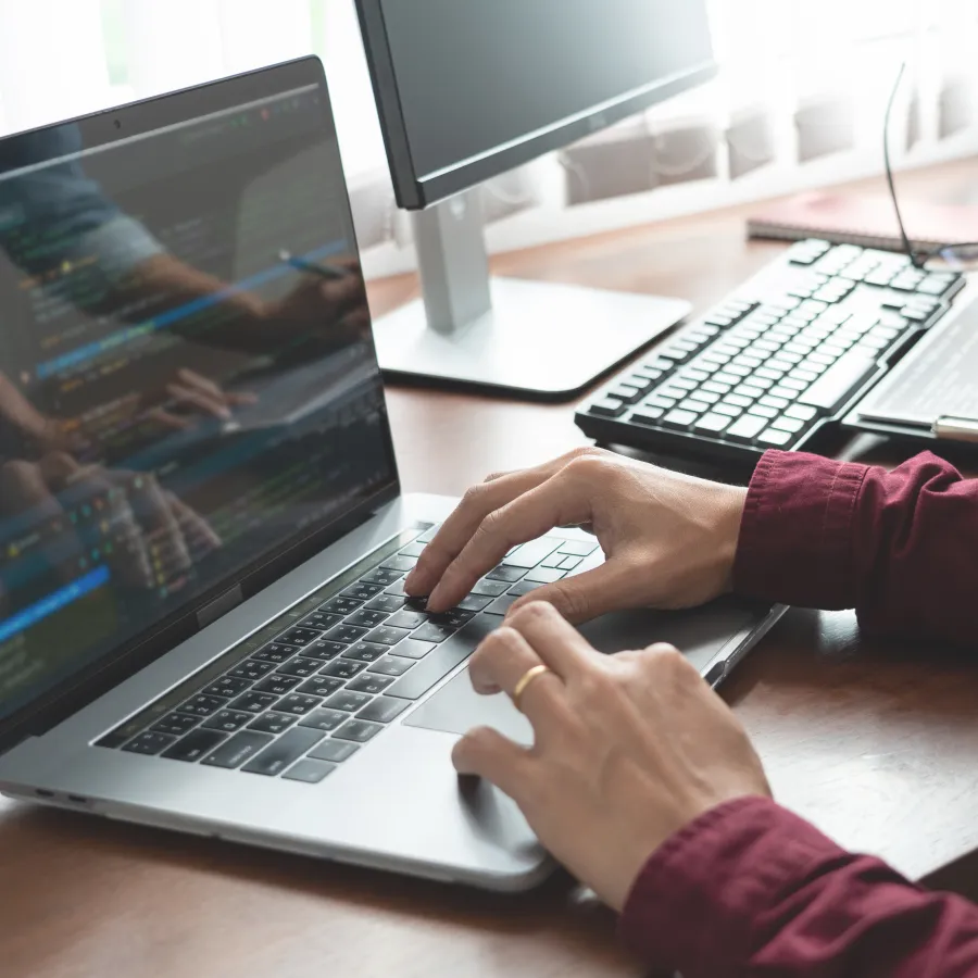 Person typing on a laptop with code on screen, another person reviewing data on a desk nearby.
