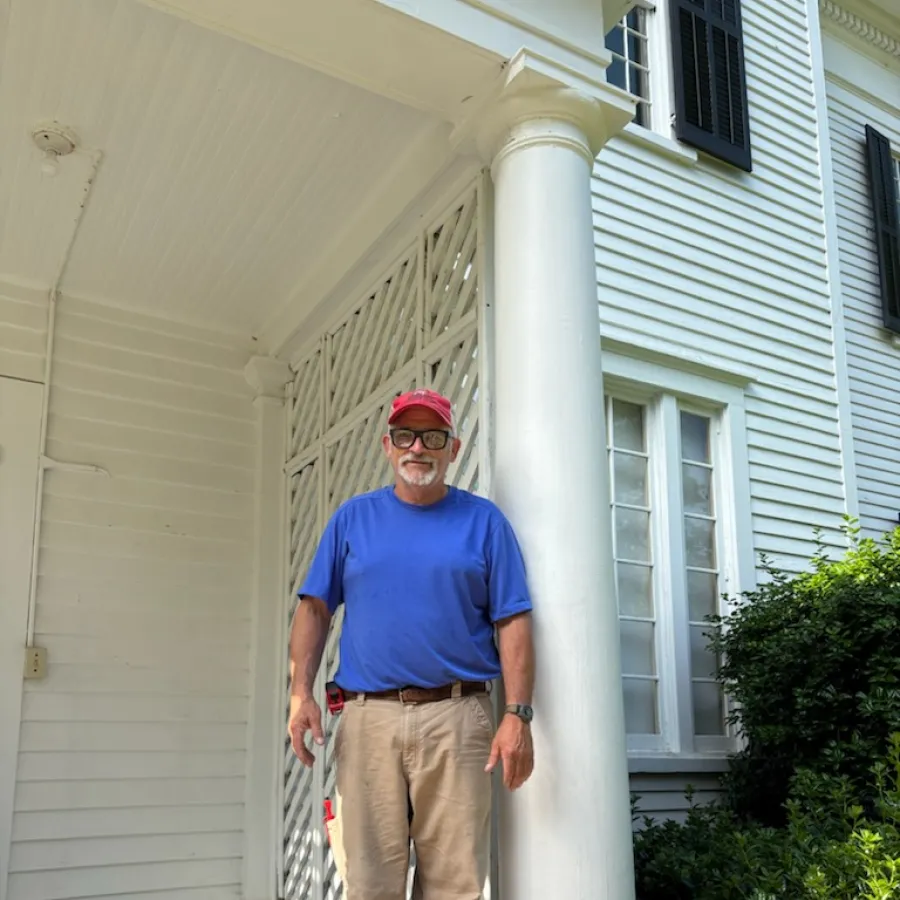 Man in blue shirt and red cap standing on a porch with white column and house exterior in background.