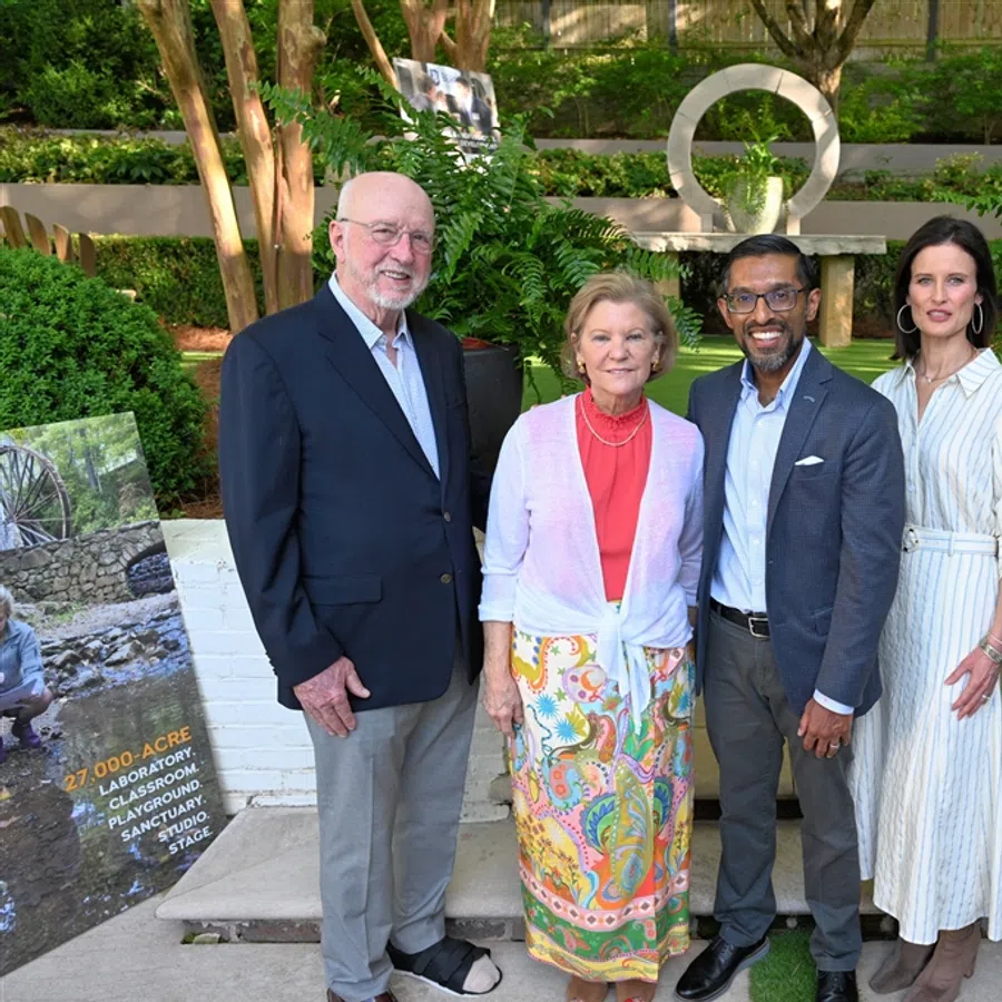Five professionally dressed people posing outdoors in a garden near a white bench and greenery during daytime.