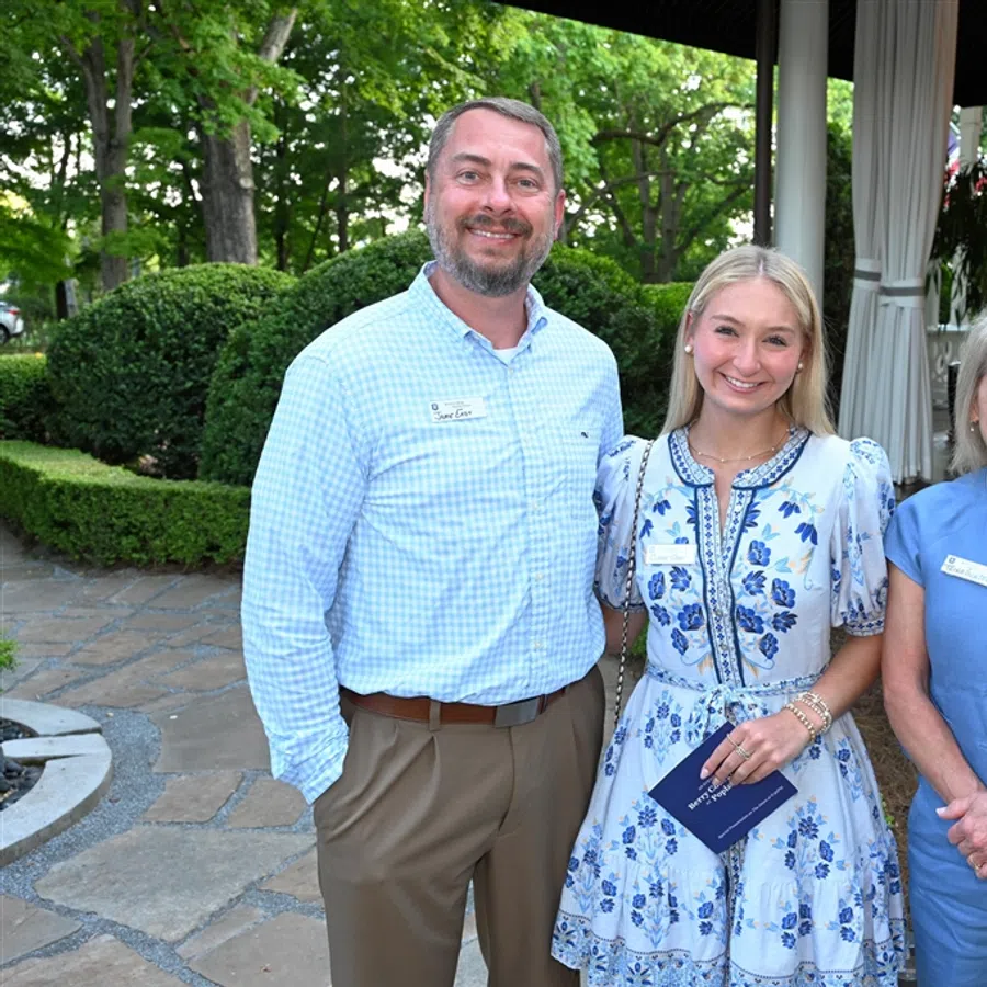 Three smiling adults standing outdoors near a garden and patio, dressed in business casual attire.