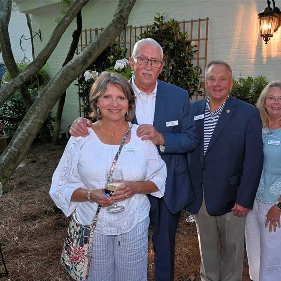 Group of five adults smiling outside a house at dusk, dressed in smart casual attire, holding drinks.