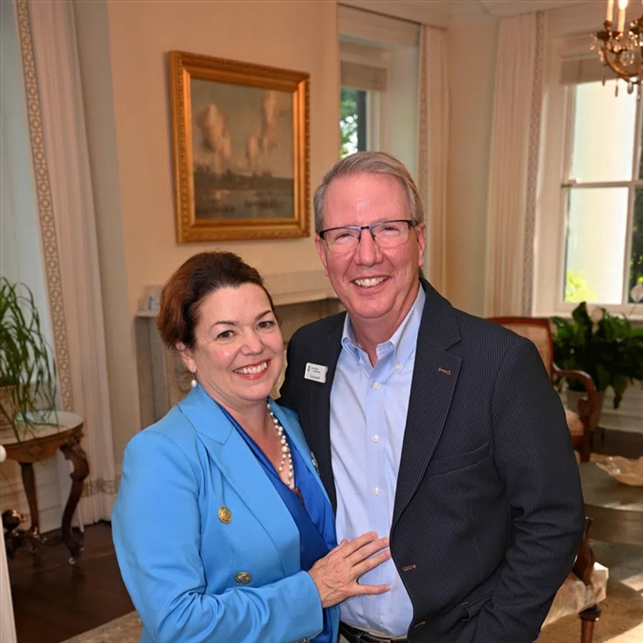 Smiling middle-aged couple in business attire poses indoors in a warmly decorated room with classic furniture and artwork.