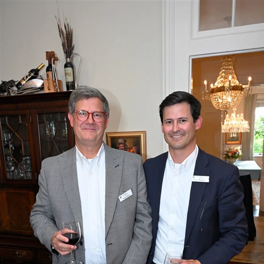 Two men in business attire holding drinks and papers, standing in a room with wood furniture and a chandelier.