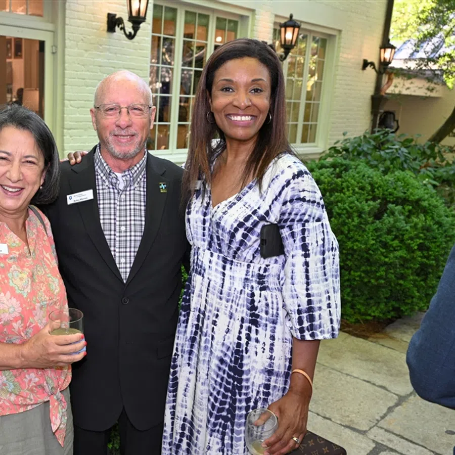 Three diverse adults smiling together outdoors at a social event near a house with greenery in the background