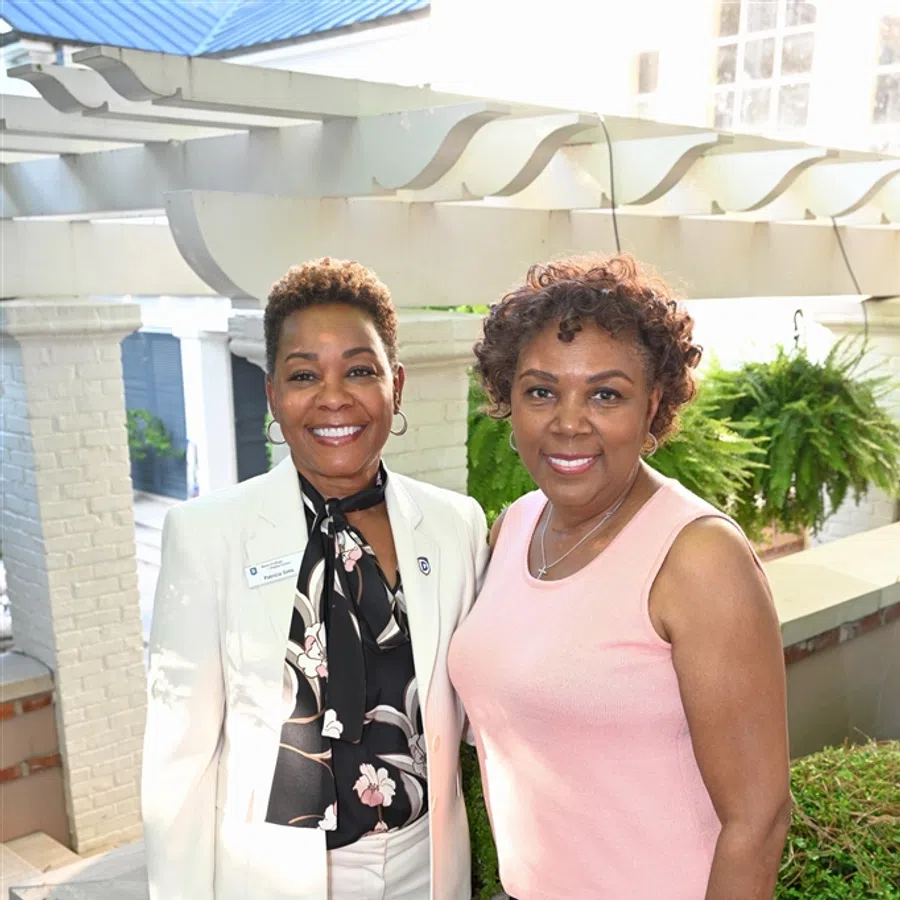 Two smiling women standing outdoors near white brick structures and green plants, dressed in business casual attire.