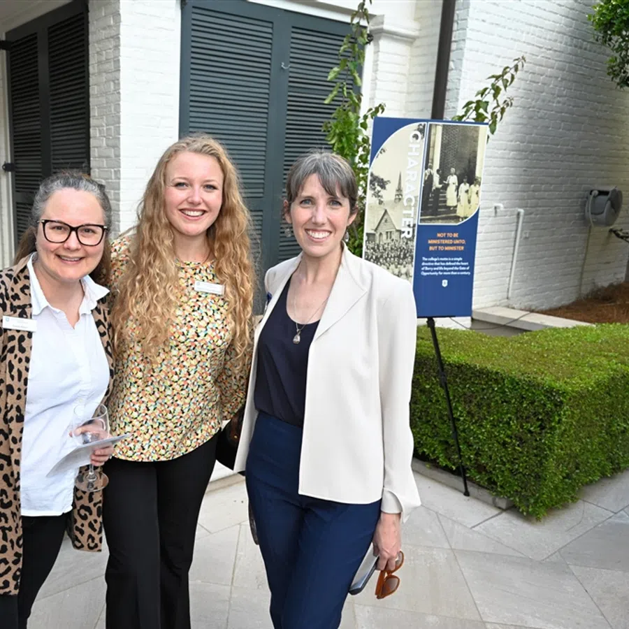 Three women smiling and posing at an outdoor event near a building entrance with guests in the background
